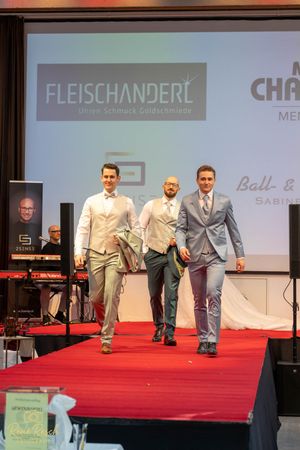 Men's formal wear presentation featuring light colored suits on a red carpet runway.