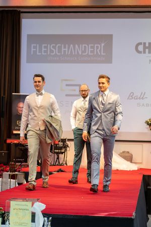 Models walking the runway in light-colored formal suits during a wedding fashion show.