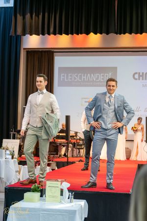 Two models showcase formal menswear on a red carpet runway with Fleischanderl branding displayed in the background.