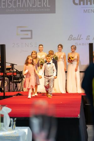Wedding party members walk the runway at a bridal fashion show featuring formal attire.