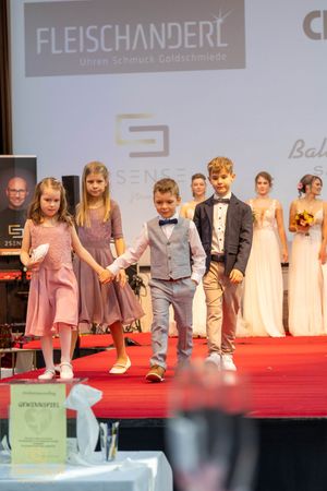 Children and adults modeling formal wear on a red carpet runway at a fashion event.