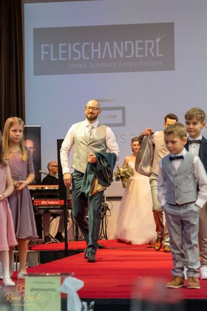 Models of various ages showcase formal wear on a red carpet runway at a Fleischanderl fashion event.