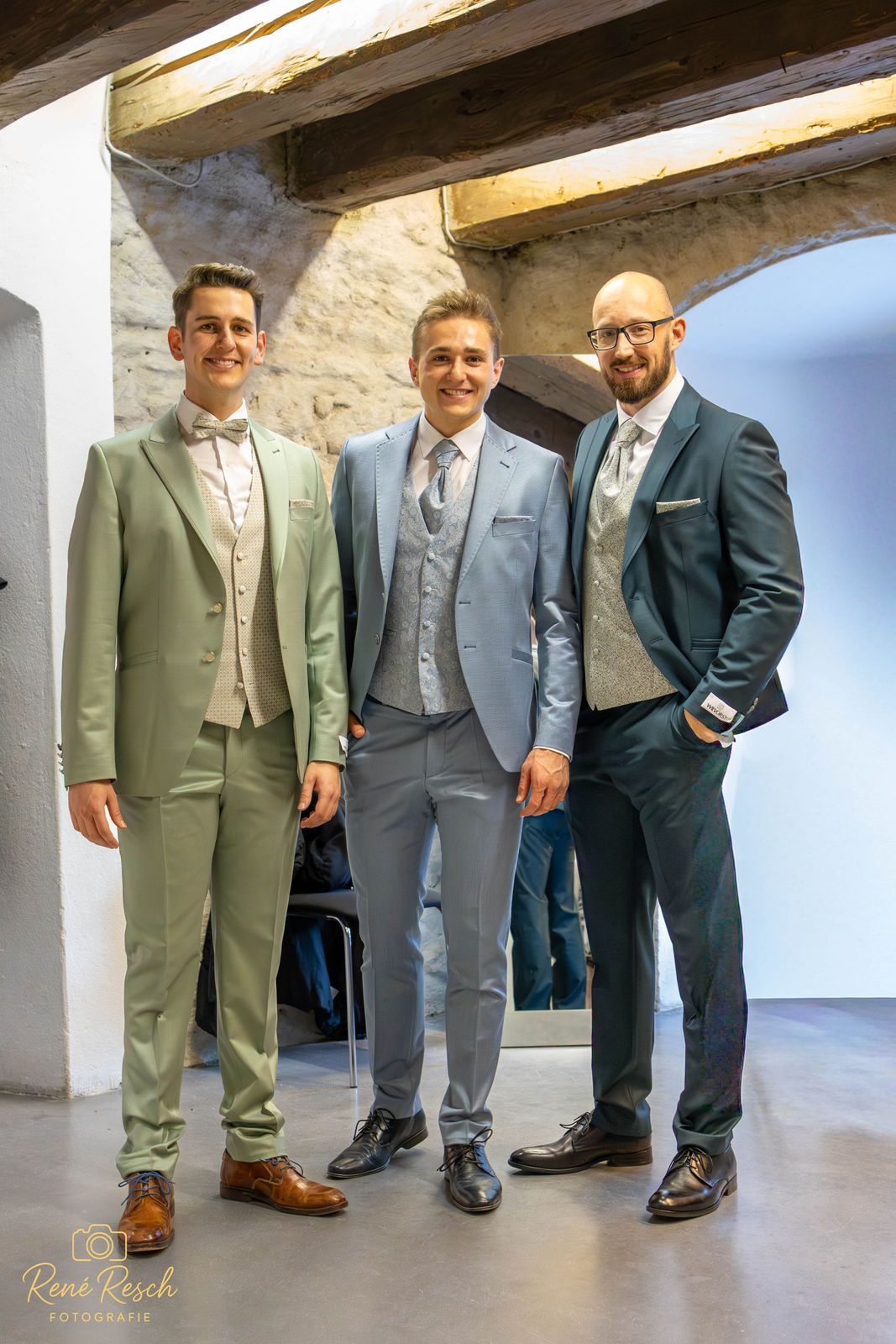 Three men pose in stylish suits against a white stone wall background.