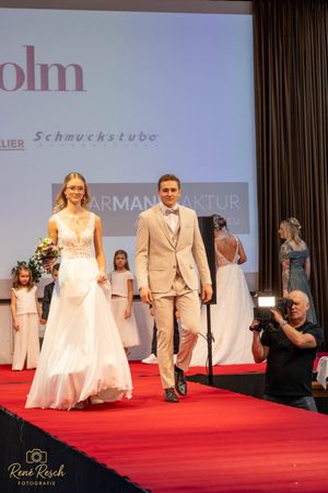 Wedding fashion show finale with models in bridal gowns and formal suits on red carpet stage.