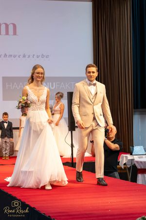Formal fashion show with models displaying wedding attire on a red carpet stage.