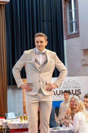 Models showcase light colored formal suits while walking on a red carpet runway.