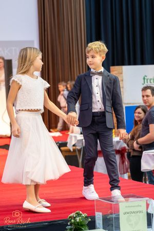 Children's fashion show displaying formal wear and wedding attire on a red carpet stage.