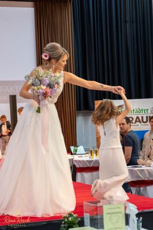 A bride in a white gown twirls with a young girl in a matching dress on a red carpet runway.