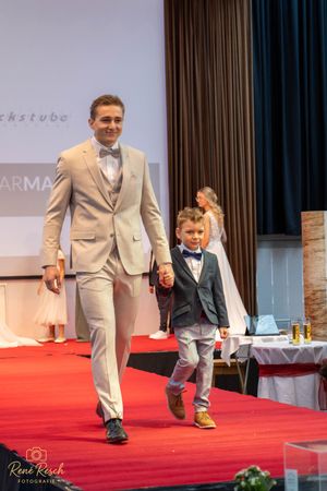Two models present formal wear while walking down a red carpet at a fashion show.