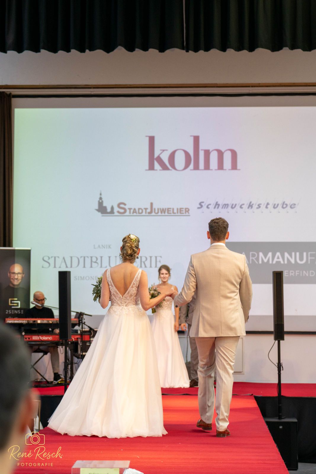 Models showcase elegant wedding attire on a red carpet runway with branding displays in the background.