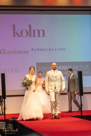 Models showcase elegant wedding attire on a red carpet runway with branded backdrop displays.