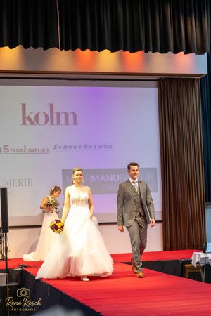 Bridal fashion show featuring white wedding gowns and formal suits on a red carpeted catwalk.