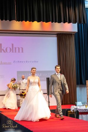 Wedding fashion show models walking down red carpet runway displaying bridal gowns and suits.