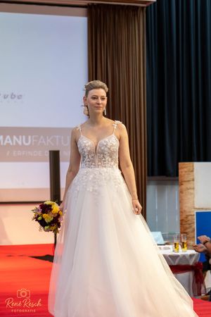 Bridal model showcasing a white lace and tulle wedding gown with bouquet on red carpet runway.