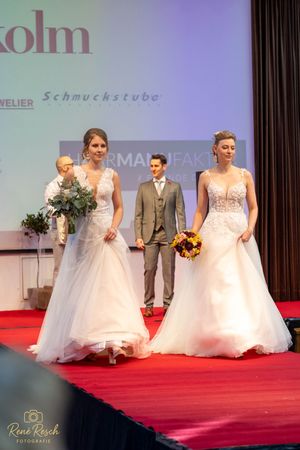 Two brides in white wedding gowns walk a red carpet runway at a bridal fashion show, holding bouquets.