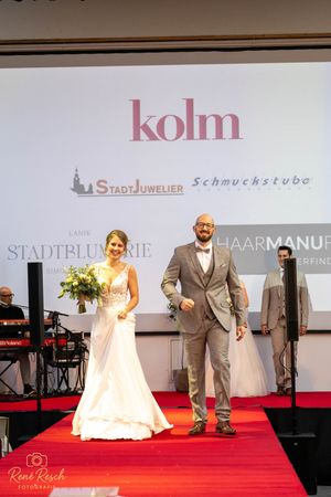 Wedding fashion show features formal gowns and suits on a red carpeted stage with sponsor logos.