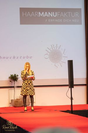 Fashion show on red carpet stage with projector screen displaying 'HAARMANUFAKTUR' text in background.