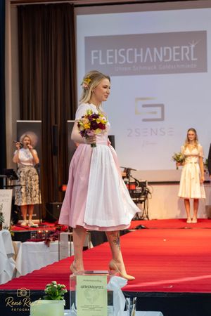 Model presents pink and white dirndl while holding floral bouquet on fashion runway.