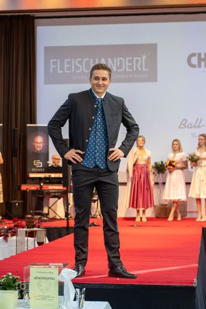 Men in dark suits showcase formal wear on red carpet runway with Fleischanderl branding.