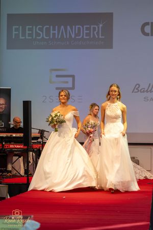 Wedding dress models lined up on stage during a bridal fashion show with Fleischanderl logo backdrop.