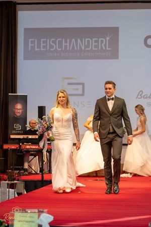 Models and groomsmen walk the runway displaying formal wedding attire at a bridal show.