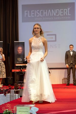 Bridal models present a collection of white wedding dresses on a red carpeted stage.