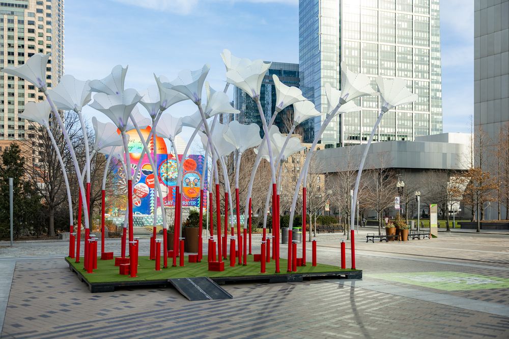 Art installation with red poles and reflective metal sculptures stands in an urban plaza between modern office buildings.