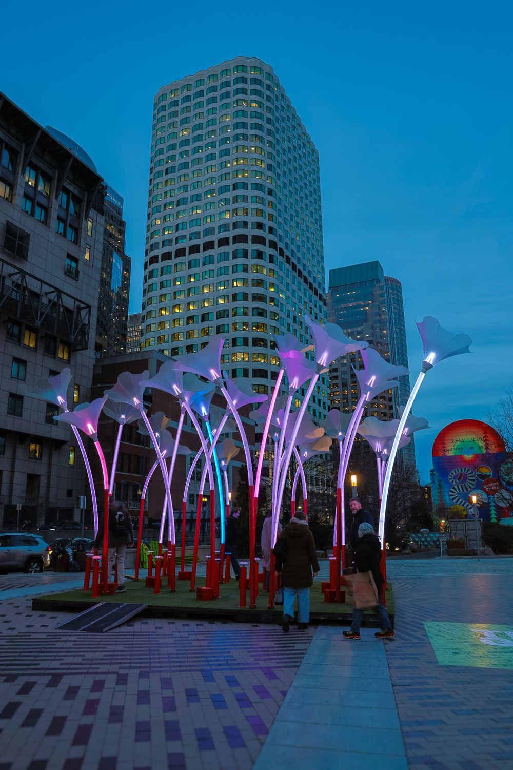 Illuminated color-changing LED light sculptures line an urban plaza at dusk with modern office buildings in the background.