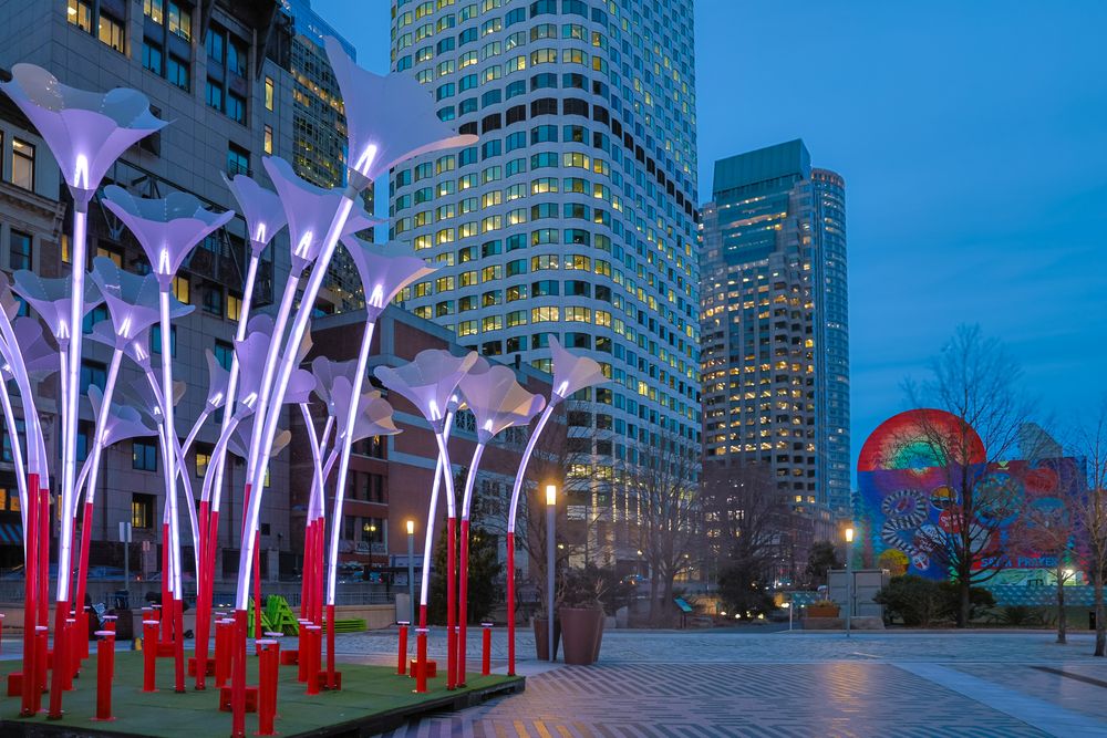 Illuminated white light poles glow against modern city buildings and blue twilight sky in an urban plaza setting.