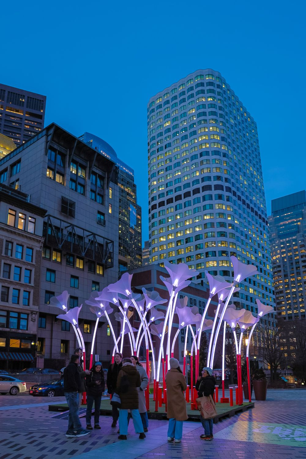 Illuminated LED light installation with glowing white stems creates an interactive art display in an urban plaza at dusk.