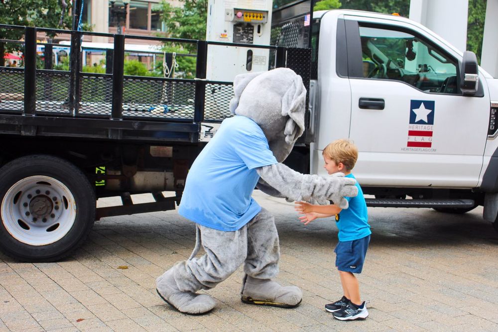 A person in a mascot costume greets a small child in front of a maintenance truck.