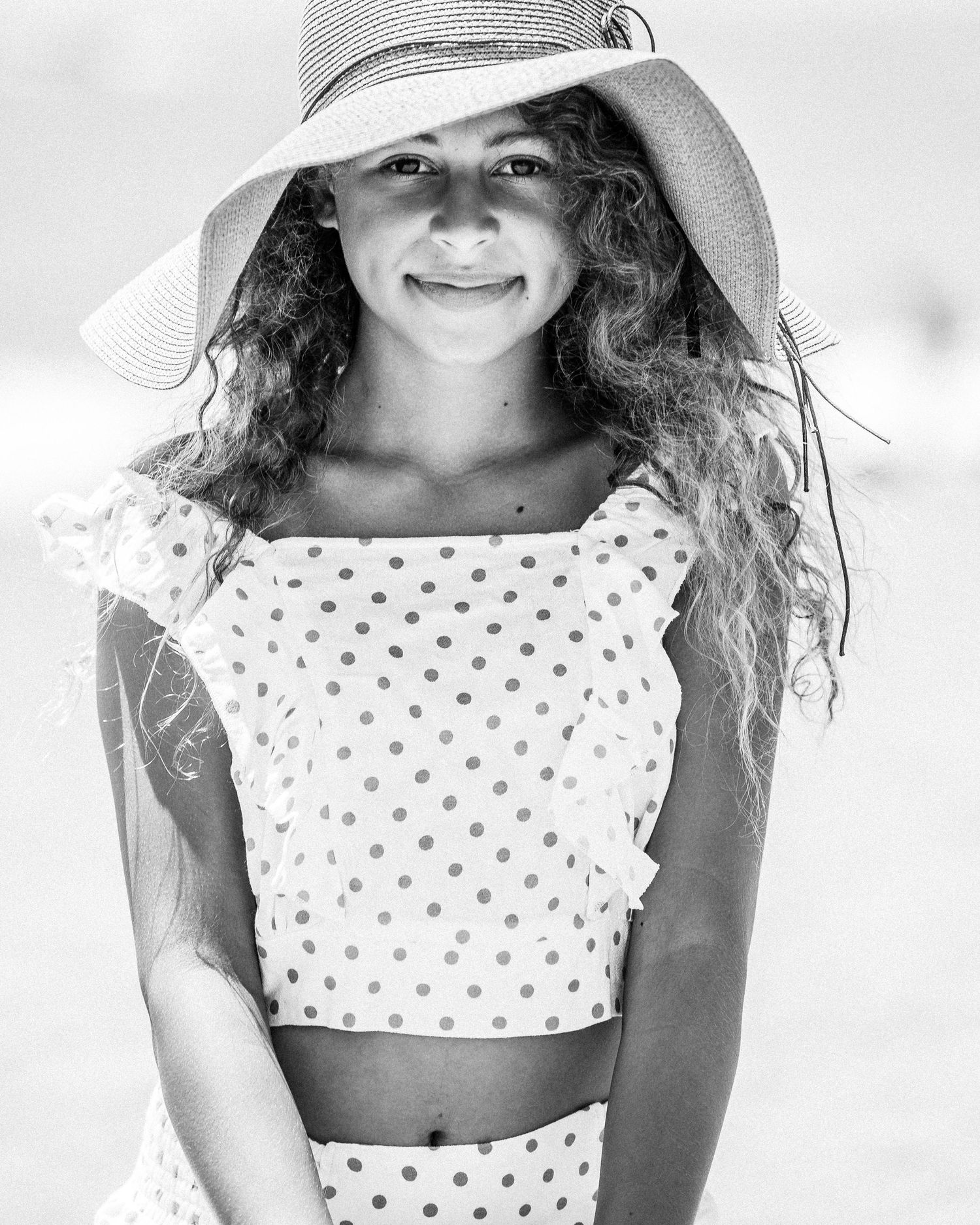 Black and white summer portrait of someone in a polka dot outfit and wide-brimmed sun hat on a bright sunny day.