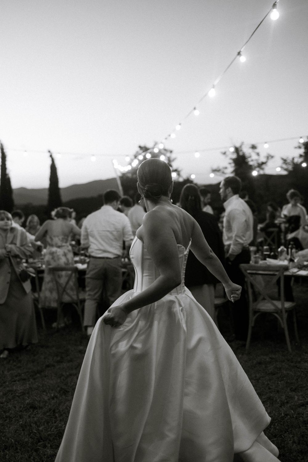 A black and white photo of an outdoor wedding reception with string lights and a person in a flowing white dress.