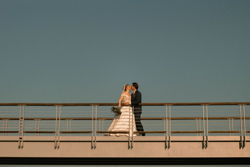 Two people in wedding attire embrace on a balcony at sunset creating a romantic silhouette against the sky.