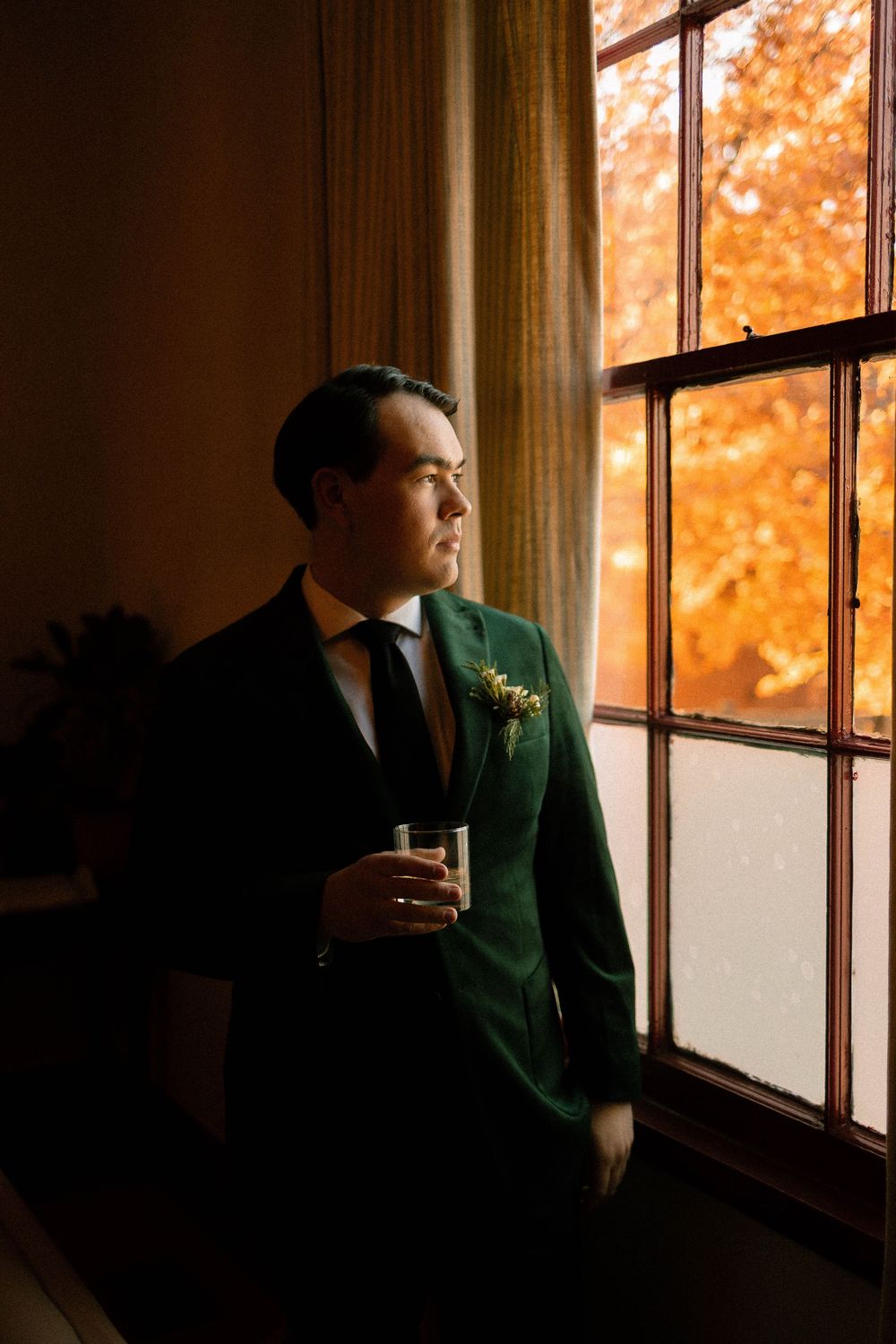 A person in a green suit gazes thoughtfully through a window with autumn foliage visible outside.