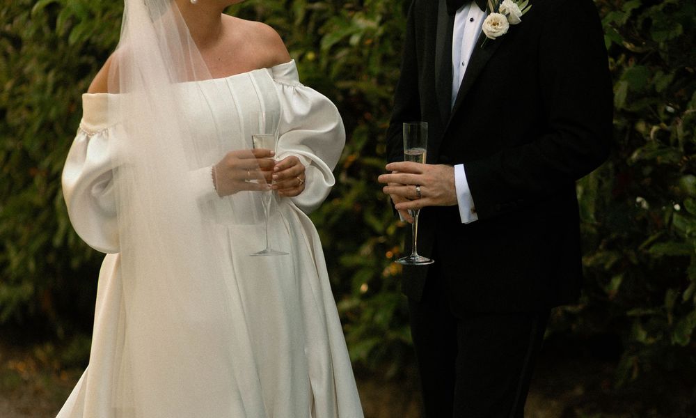 Bride in flowing off-shoulder white gown and groom in tuxedo hold champagne glasses during outdoor wedding celebration.