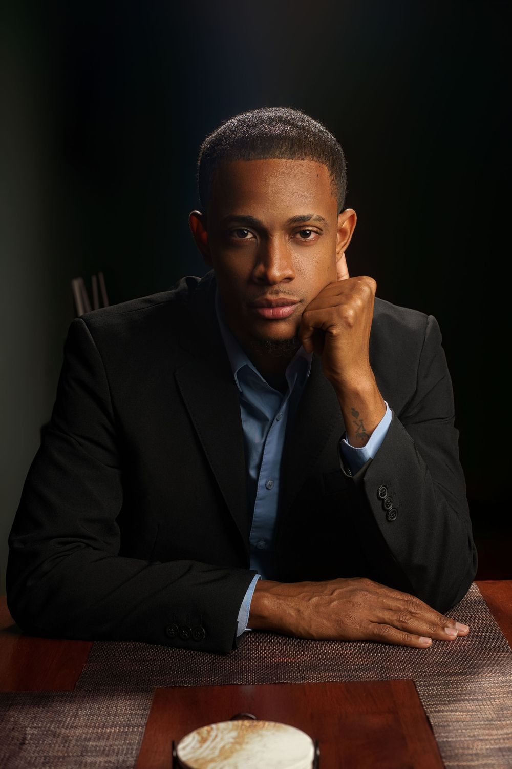 Professional portrait series showing someone in a dark suit jacket resting against a wooden table.