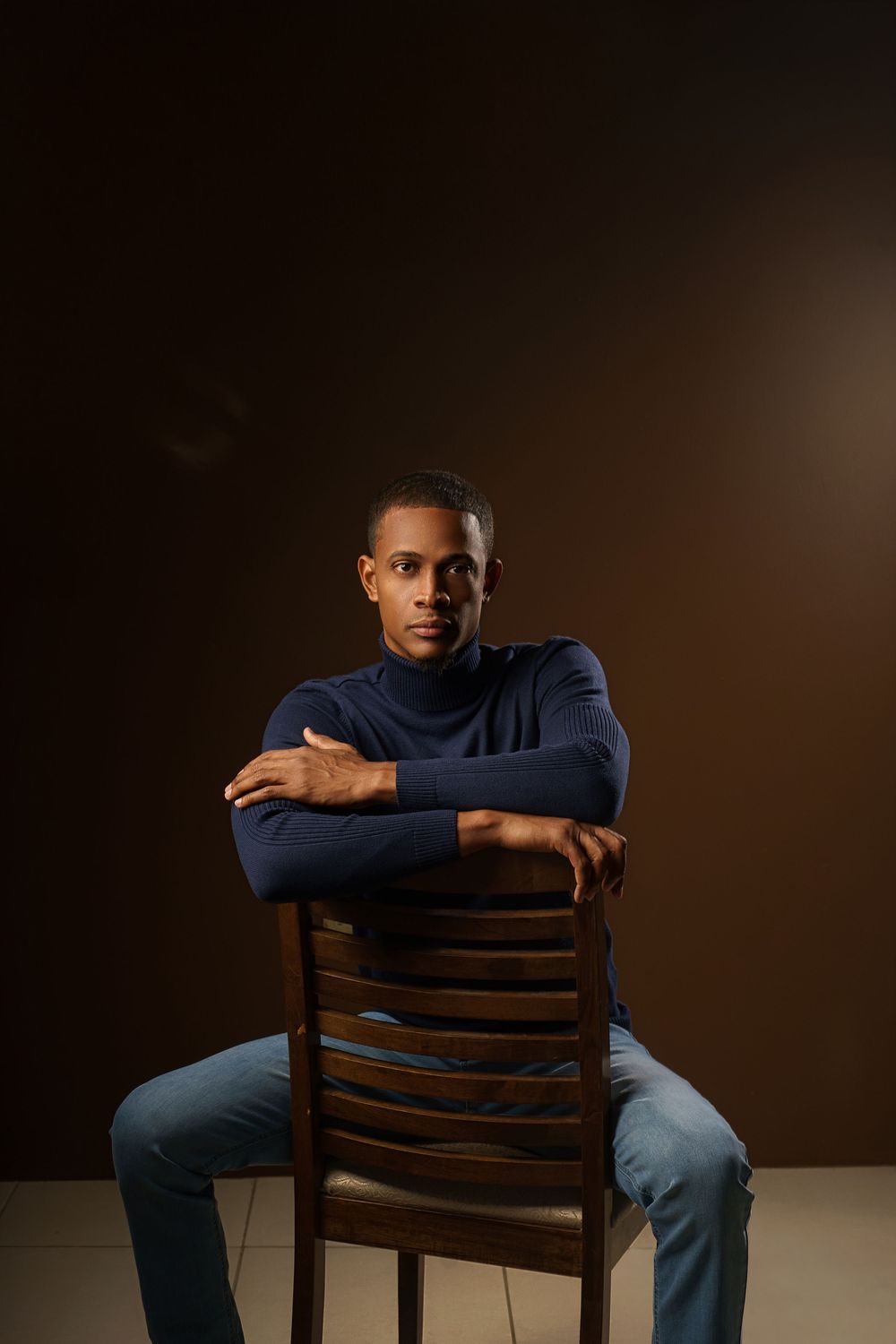 Moody studio portraits of someone sitting backwards on a wooden chair against a dark background.