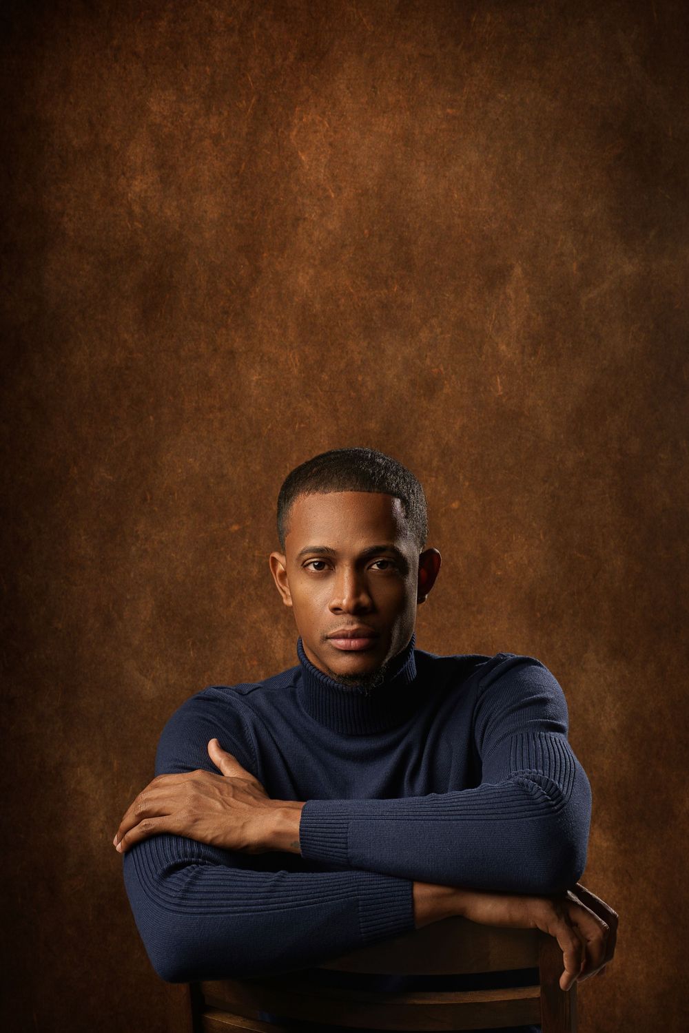Professional studio portrait against a brown textured background with dramatic lighting and navy blue sweater.