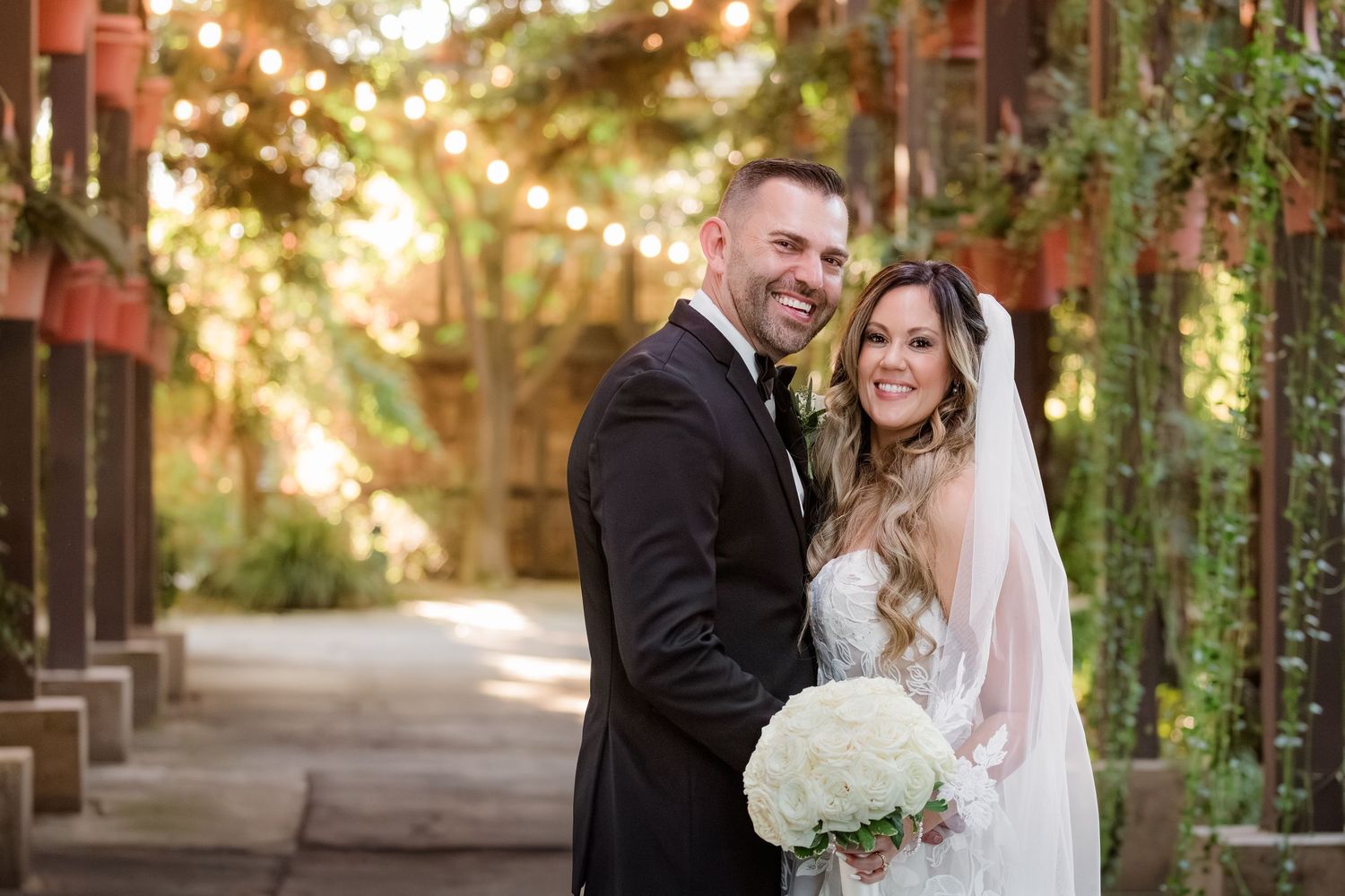 A bride and groom share joyful moments together in a sunlit garden walkway with white flowers and greenery.