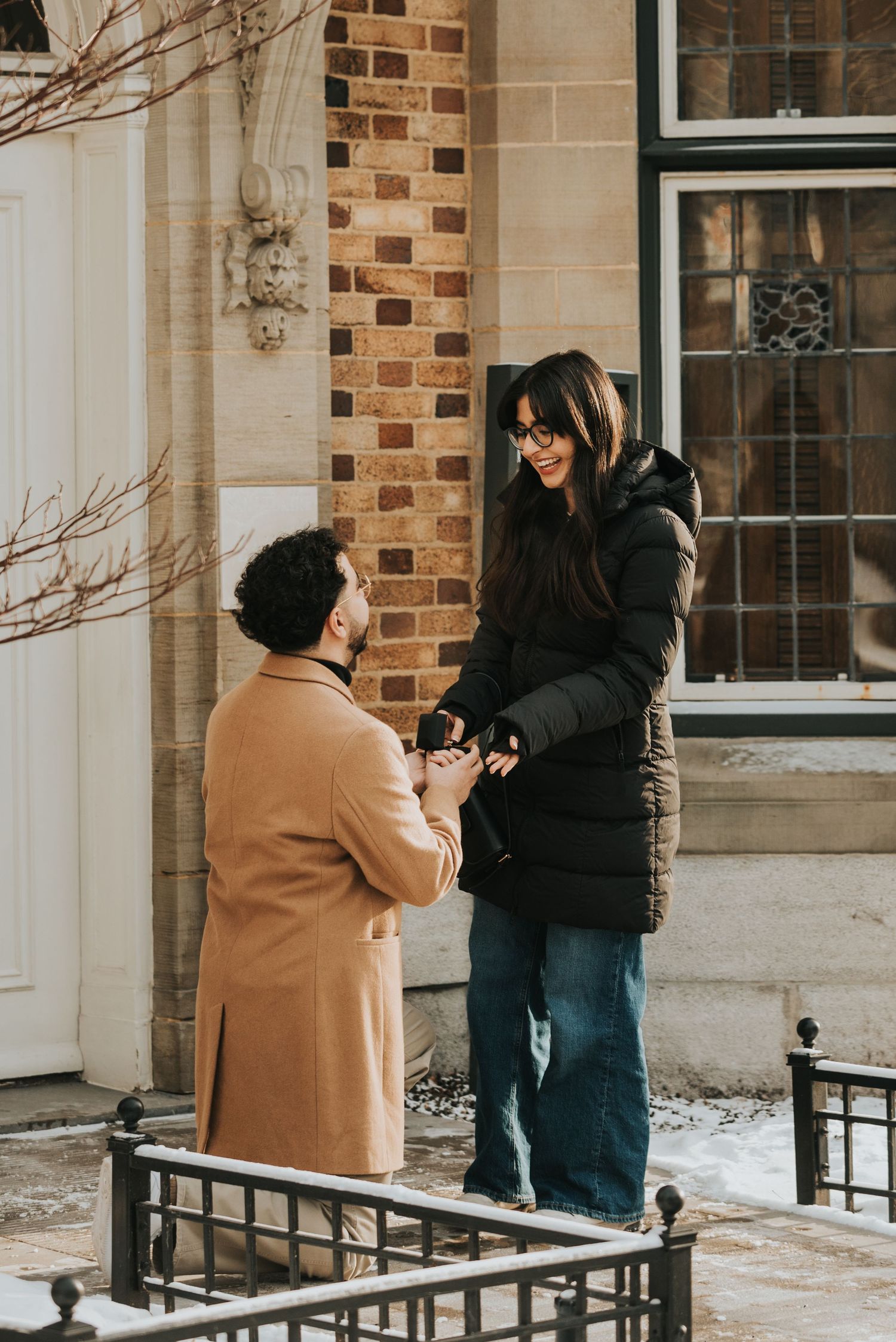 A couple stands together in winter outside a brick building with white columns during a proposal moment.