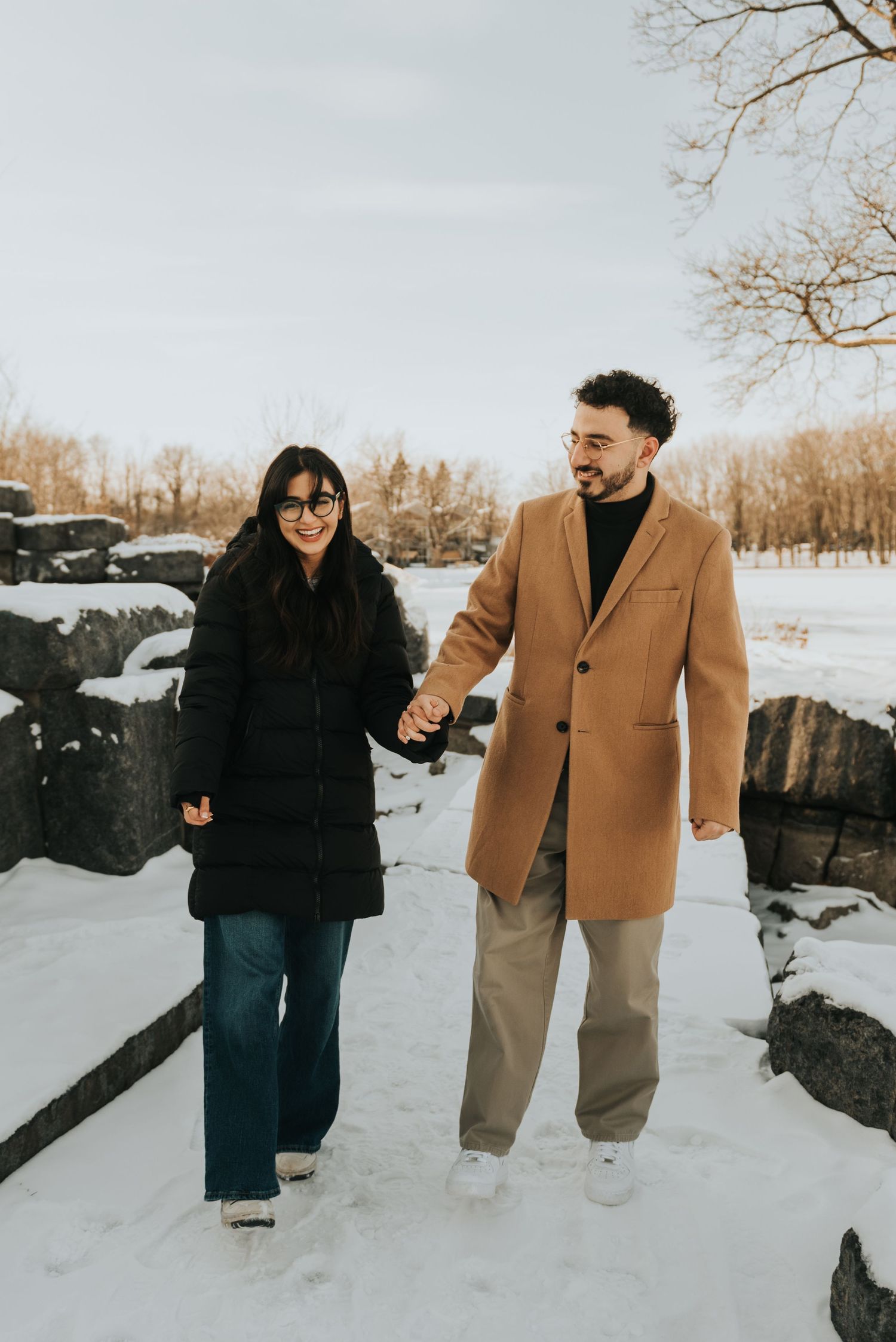 A couple walks hand in hand through a snowy winter landscape wearing a black coat and camel coat.