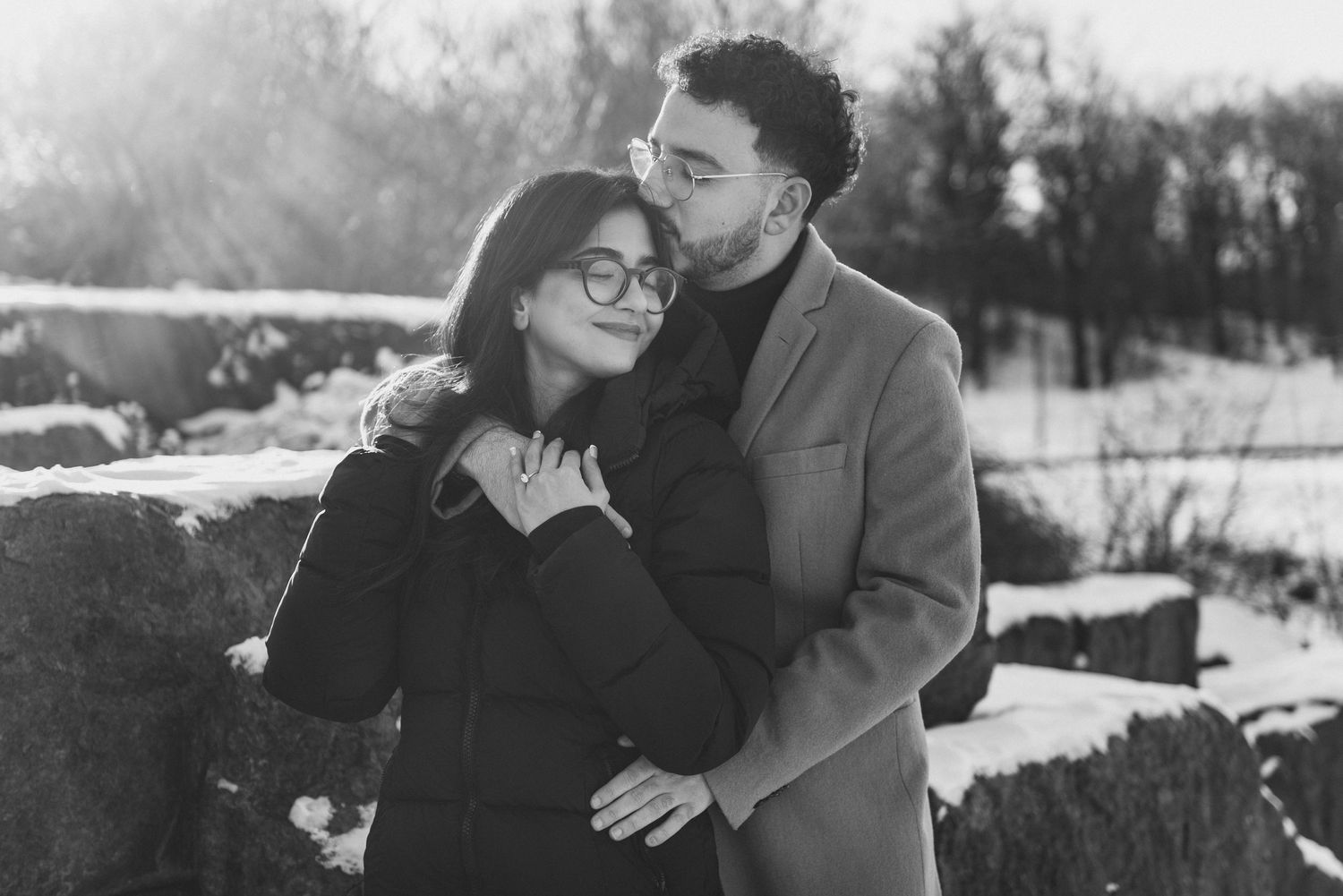 A romantic black and white winter portrait of a couple embracing outdoors in the snow with sunlight behind them.