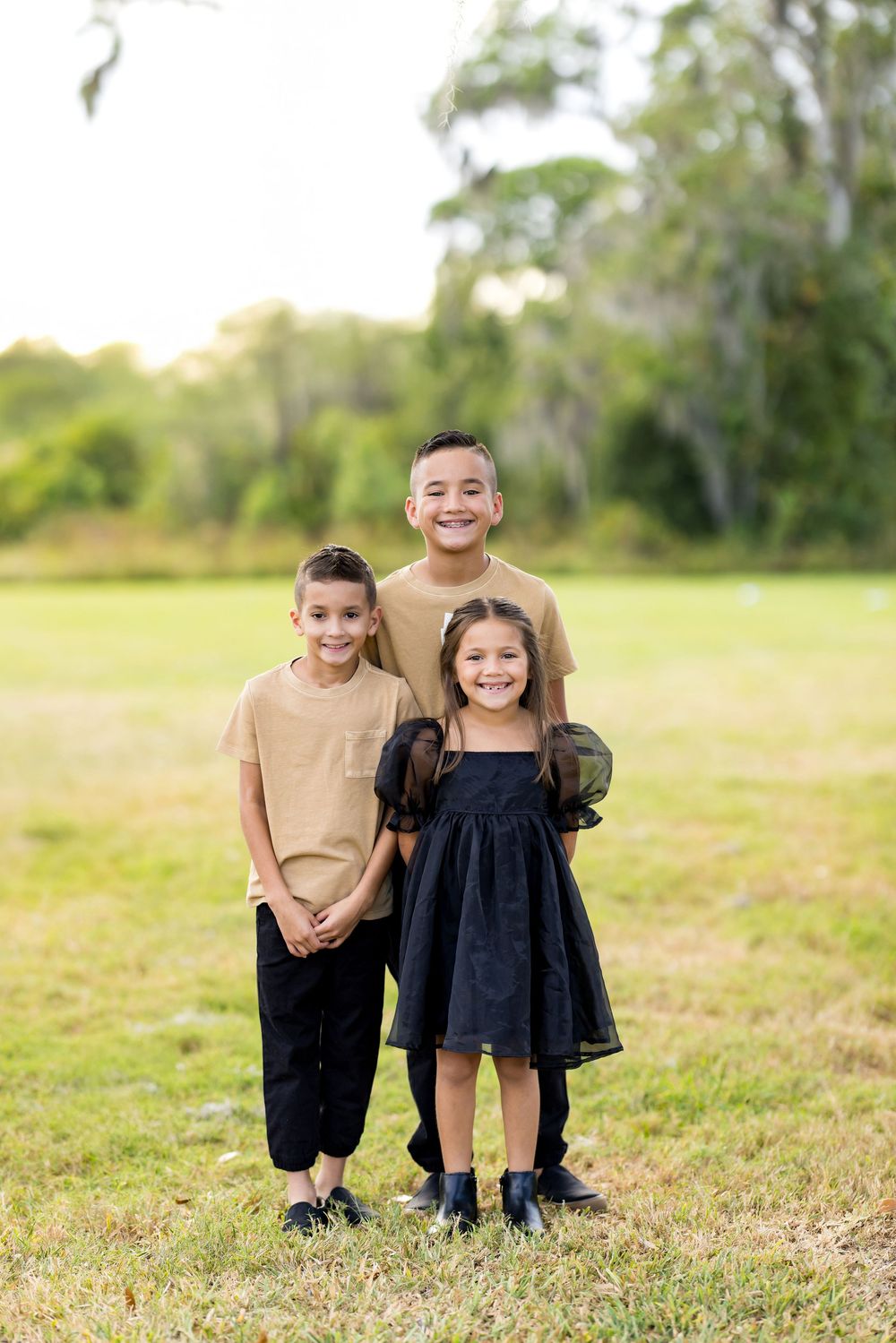 Three children stand together smiling in a grassy field with trees in the background during golden hour.