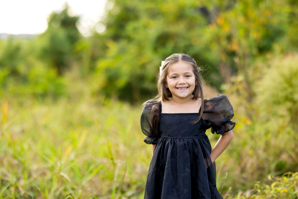 A young person in a black dress poses outdoors in a field of tall grass and wildflowers on a sunny day.