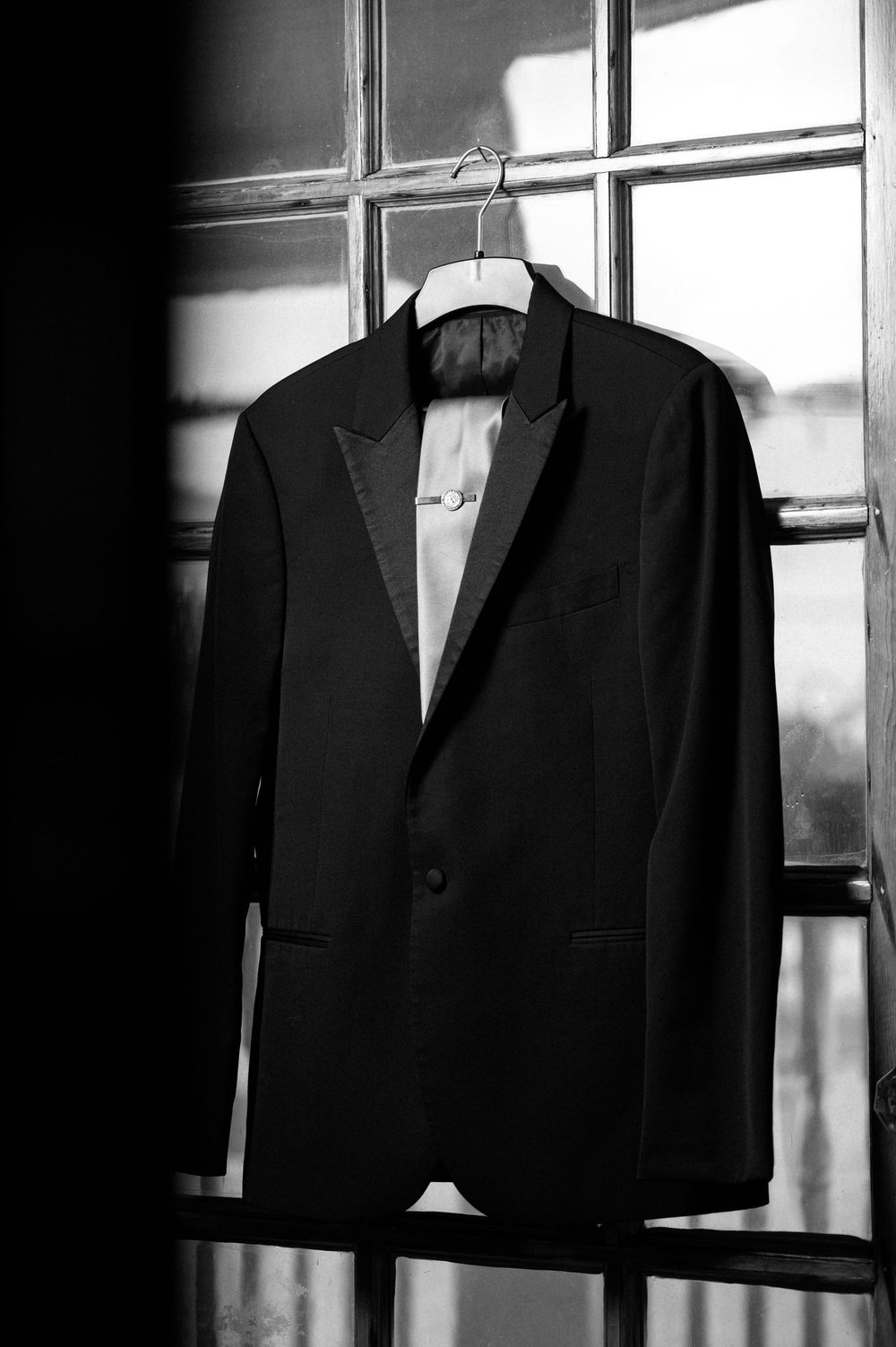 A black tuxedo jacket hangs on display against a window in dramatic black and white lighting.