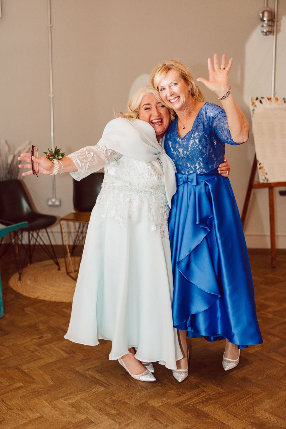Two women in elegant dresses, one in white and one in royal blue, dance together on a parquet floor at a celebration.