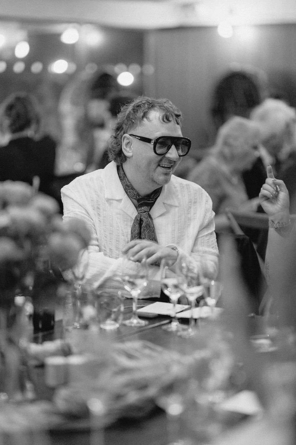 A guest in sunglasses and white jacket sits at a formal dinner table during a celebration event.