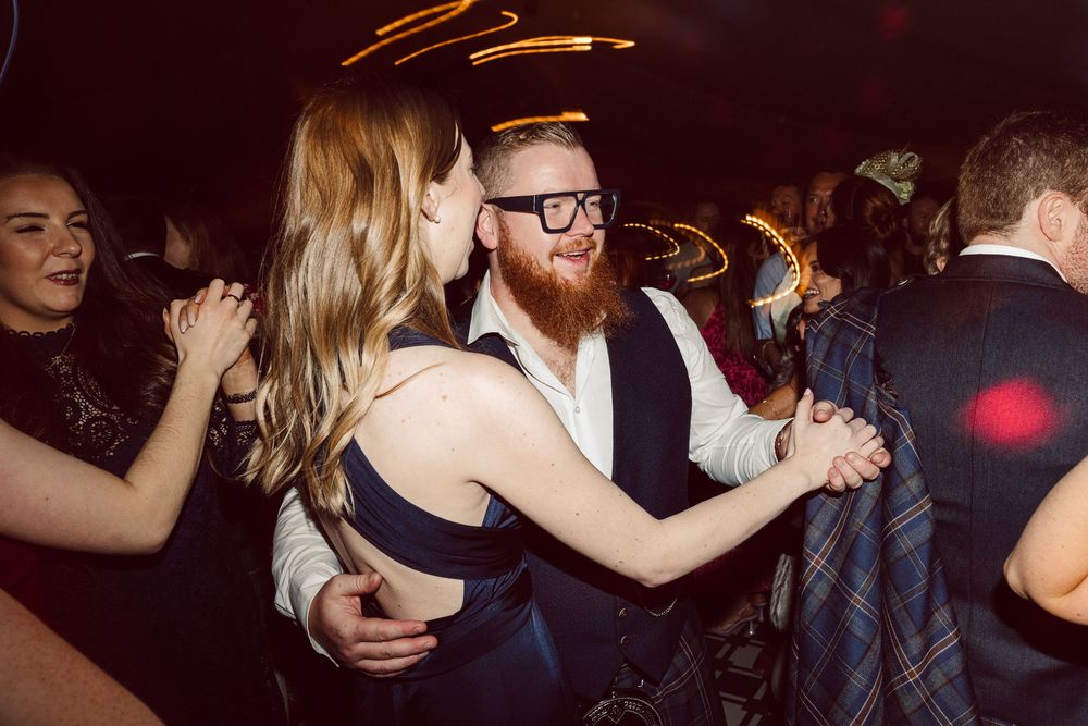 Wedding guests dance and celebrate under dim lighting at an evening reception.