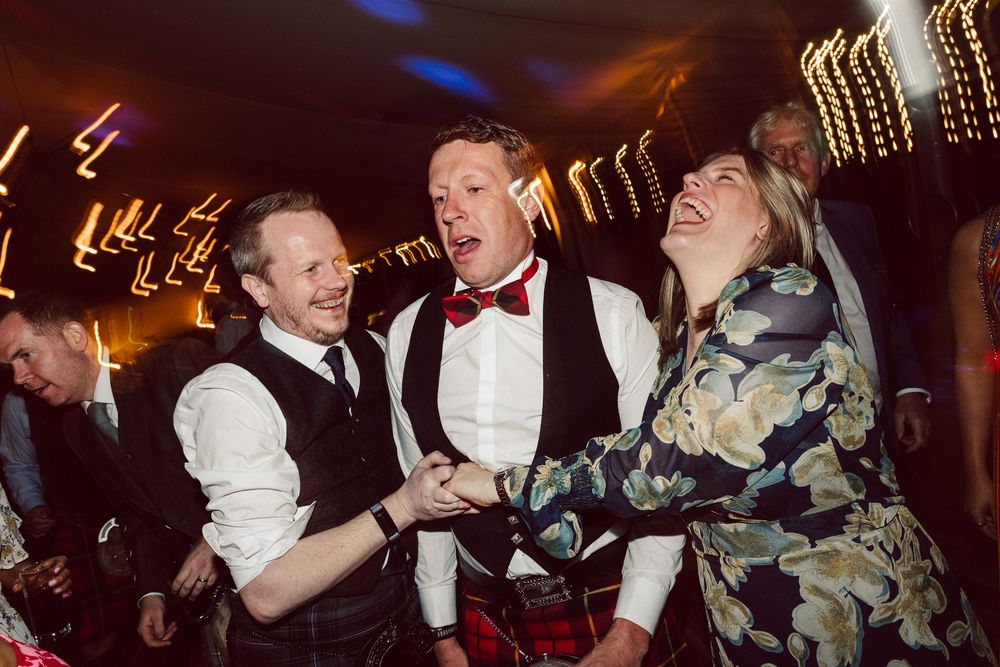 Party guests dance and laugh together at a lively wedding reception with blurred lights in the background.